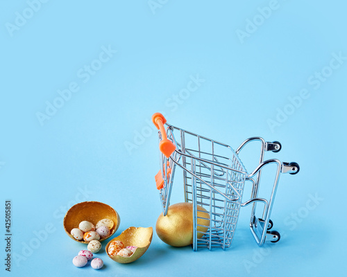 Easter sale concept. Golden easter egg fell from small shopping cart and shattered on blue background photo