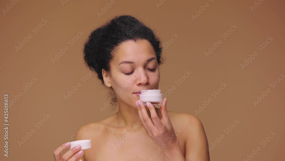 Beauty portrait of young African American woman twisting off a jar of cream and enjoying its aroma. Black female model posing on brown studio background. Slow motion ready, 4K at 59.94fps. Beauty portrait of young African American woman twisting off a jar of cream and enjoying its aroma. Black female model posing on brown studio background. Slow motion ready, 4K at 59.94fps.