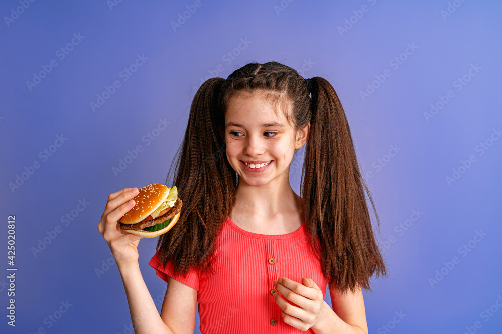 happy, cheerful teenage girl with two ponytails is holding a delicious hamburger on a purple background. Positive child loves fast food.fast food concept. Copy space happy, cheerful teenage girl with two ponytails is holding a delicious hamburger on a purple background. Positive child loves fast food.fast food concept. Copy space