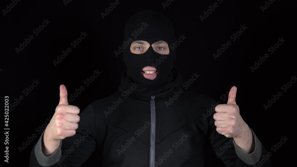 A man in a balaclava mask shows his approval mark with his hands. The thug raised his hands and showed his thumbs up against a black background. A man in a balaclava mask shows his approval mark with his hands. The thug raised his hands and showed his thumbs up against a black background.