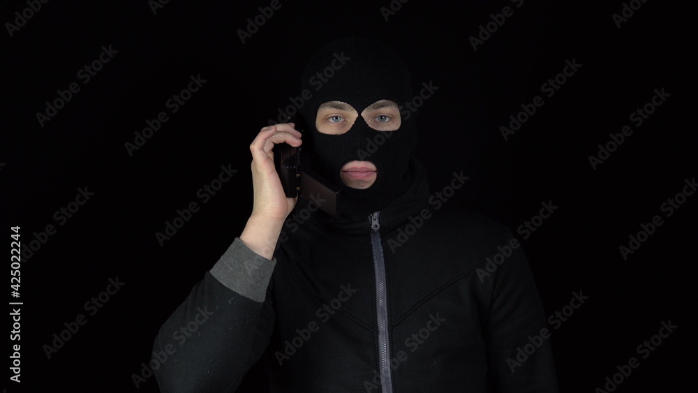 A man in a balaclava mask with a phone from the 90s. The bandit is talking on the phone. On a black background. A man in a balaclava mask with a phone from the 90s. The bandit is talking on the phone. On a black background.