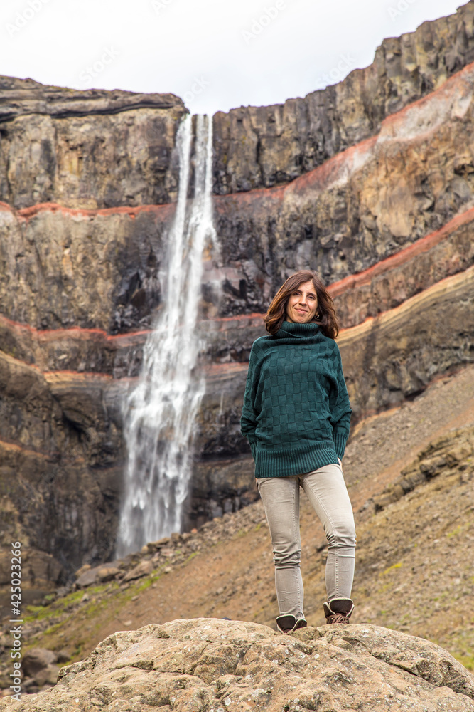 Hengifoss, Iceland »; August 2017: A young woman with a green sweater under the Hengifoss waterfall sitting on a stone.