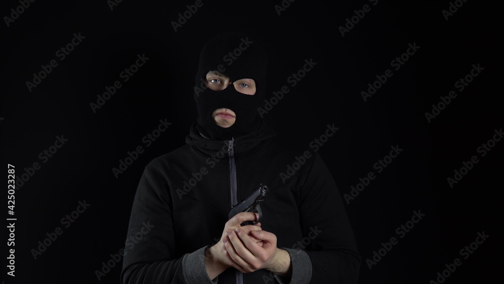 A man in a balaclava mask stands with a gun. The bandit reloaded the gun and stands on a black background. A man in a balaclava mask stands with a gun. The bandit reloaded the gun and stands on a black background.