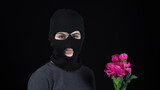 A Woman in a balaclava mask is standing with flowers. Bandit sniffs a bouquet of pink flowers on a black background.