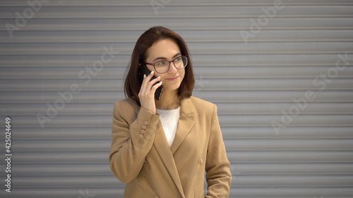 A young woman stands on a gray background and speaks on the phone.