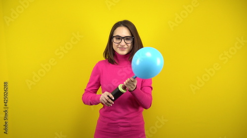 A young woman inflates a blue balloon with a pump on a yellow background. Girl in a pink turtleneck and glasses. © Vital9c