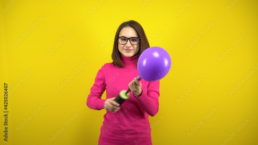 A young woman inflates a purple balloon with a pump on a yellow background. Girl in a pink turtleneck and glasses. A young woman inflates a purple balloon with a pump on a yellow background. Girl in a pink turtleneck and glasses.