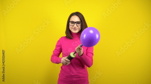 A young woman inflates a purple balloon with a pump on a yellow background. Girl in a pink turtleneck and glasses. © Vital9c