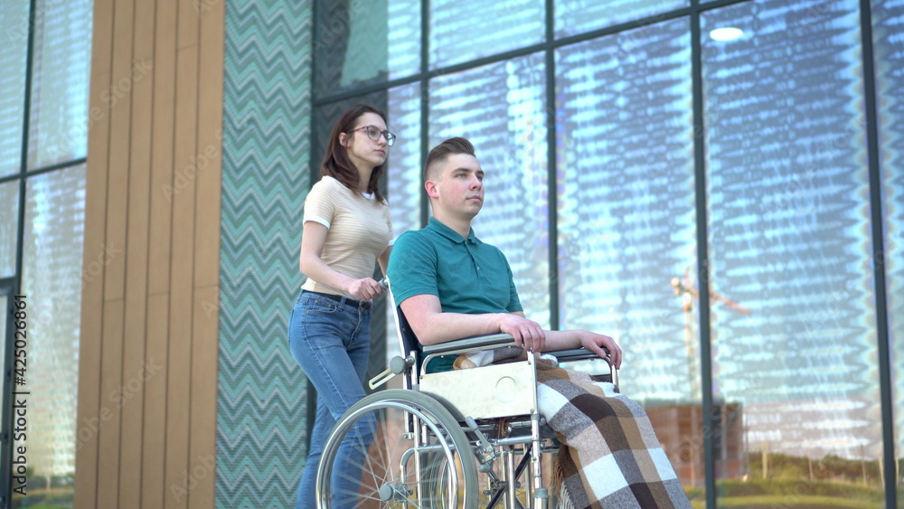A young woman is carrying a young man in a wheelchair. An assistant carries a disabled person in a wheelchair along the street. Special transport for the disabled. A young woman is carrying a young man in a wheelchair. An assistant carries a disabled person in a wheelchair along the street. Special transport for the disabled.