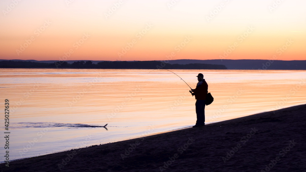 A man caught a fish. A silhouette of a man fishing at sunrise by the river. The yellow sun rises from the horizon.