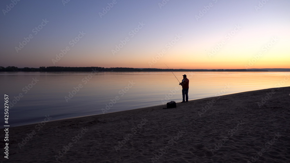 A man is fishing at sunrise by the river. The yellow sun rises from the horizon. Silhouette of a fisherman.