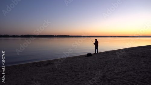 A man is fishing at sunrise by the river. The yellow sun rises from the horizon. Silhouette of a fisherman.