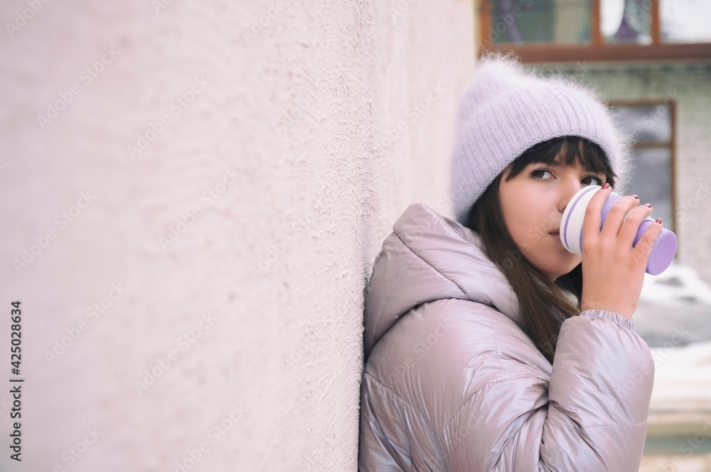 young girl dressed in a purple hat and a pink jacket stands against the backdrop of the wall and drinks coffee from an ecological reusable cup young girl dressed in a purple hat and a pink jacket stands against the backdrop of the wall and drinks coffee from an ecological reusable cup