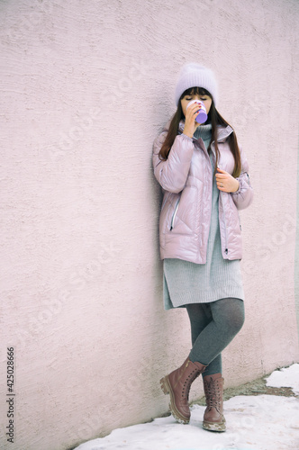 young girl dressed in a purple hat and a pink jacket stands against the backdrop of the wall and drinks coffee from an ecological reusable cup