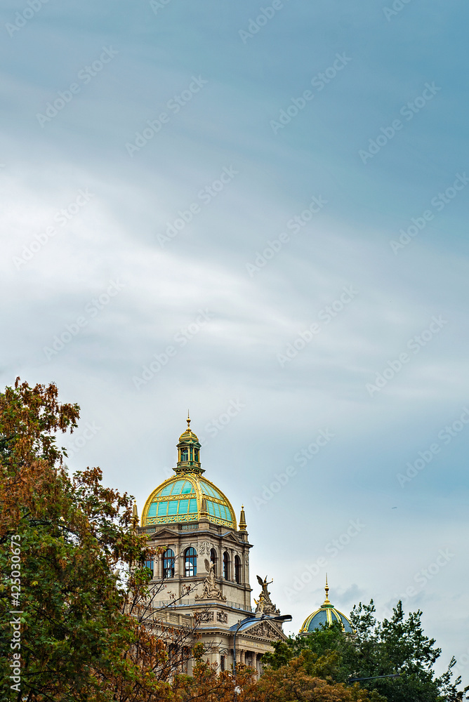 Colored dome with gilding of the National Museum in Prague in the distance against the blue sky and autumn yellow and green leaves on the trees. Statues on the facade. Ancient building in the old town