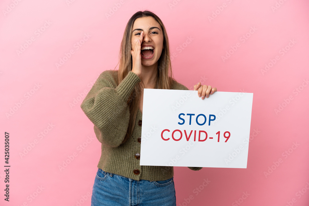 Young caucasian woman isolated on pink background holding a placard with text Stop Covid 19 and shouting Young caucasian woman isolated on pink background holding a placard with text Stop Covid 19 and shouting
