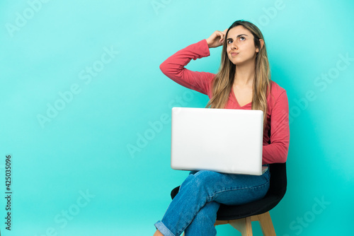 Young caucasian woman sitting on a chair with her pc isolated on blue background having doubts and with confuse face expression © luismolinero