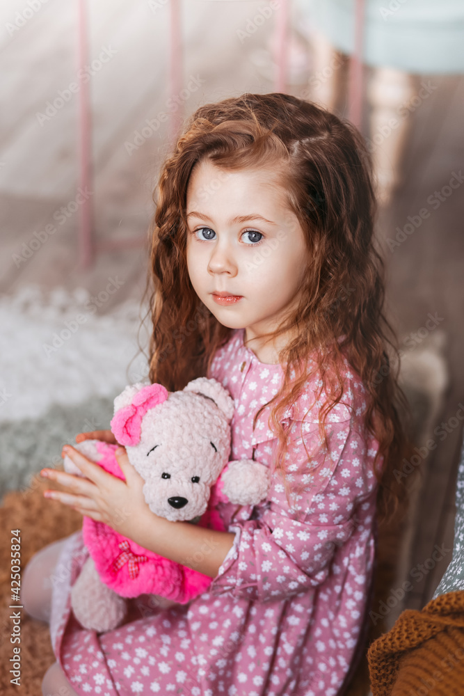 A beautiful little girl hugs a soft toy bear in a light room.