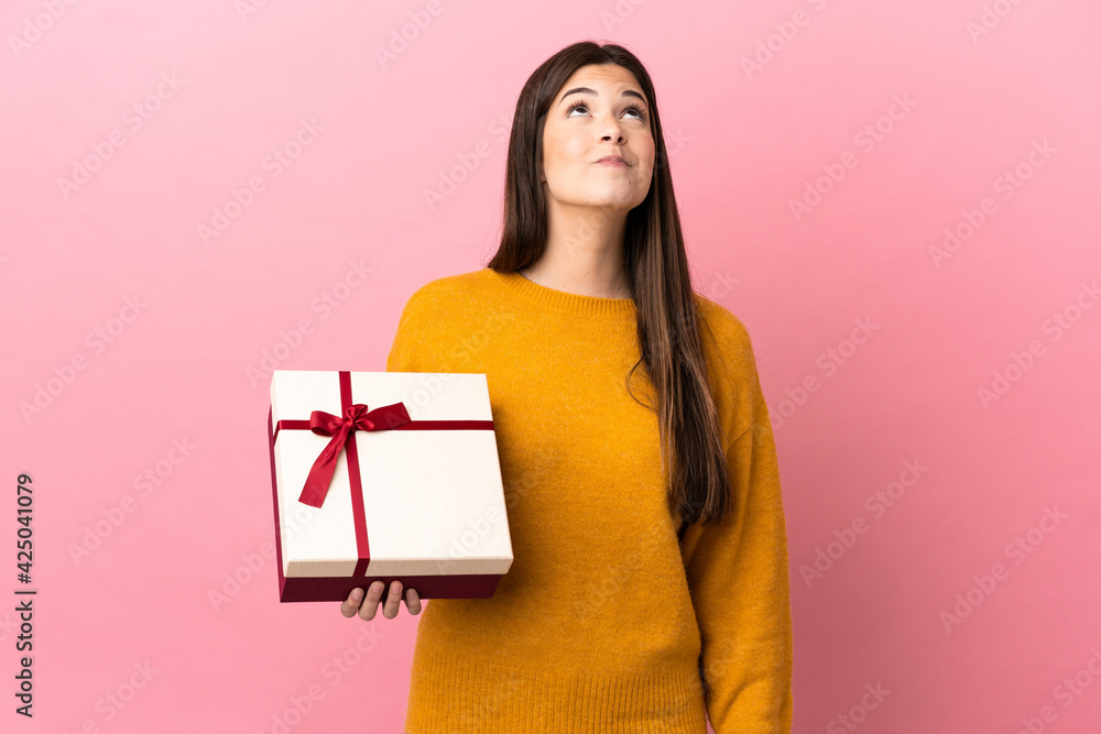 Teenager Brazilian girl holding a gift over isolated pink background and looking up Teenager Brazilian girl holding a gift over isolated pink background and looking up
