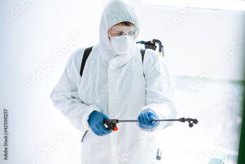 Man in Hazmat suit, protective gloves and goggles use sprayer equipment disinfect the entrance of a residential building. Covid-2019.