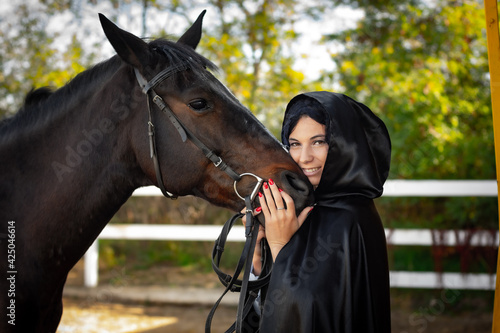 A girl in a black cloak hugs the muzzle of a horse against the background of trees and a fence
