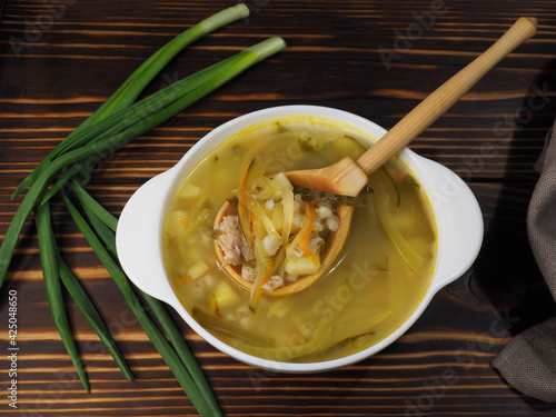 Seasonal hot soup with pickles and pearl barley in a white bowl, sour cream, spoon on a wooden background, flat layout. Traditional Russian cuisine, meat broth with vegetables for lunch photo