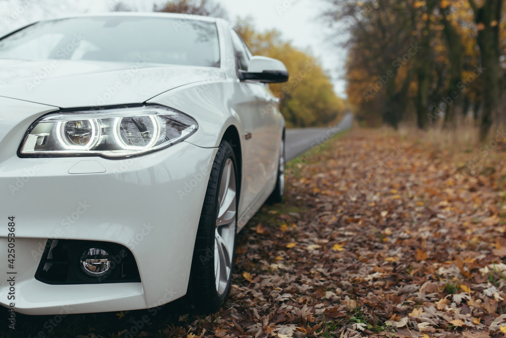 Close-up photo, part of a white premium car standing in the road in autumn, part of a car headlight and wheels