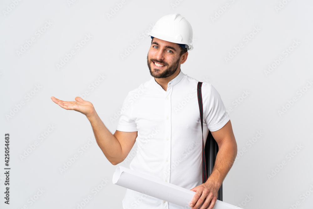 Young architect man with helmet and holding blueprints isolated on white background extending hands to the side for inviting to come Young architect man with helmet and holding blueprints isolated on white background extending hands to the side for inviting to come