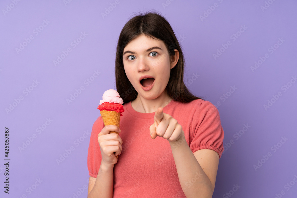 Young Ukrainian teenager girl holding a cornet ice cream over isolated purple background surprised and pointing front Young Ukrainian teenager girl holding a cornet ice cream over isolated purple background surprised and pointing front