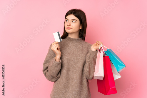 Young Ukrainian teenager girl over isolated pink background holding shopping bags and a credit card and thinking