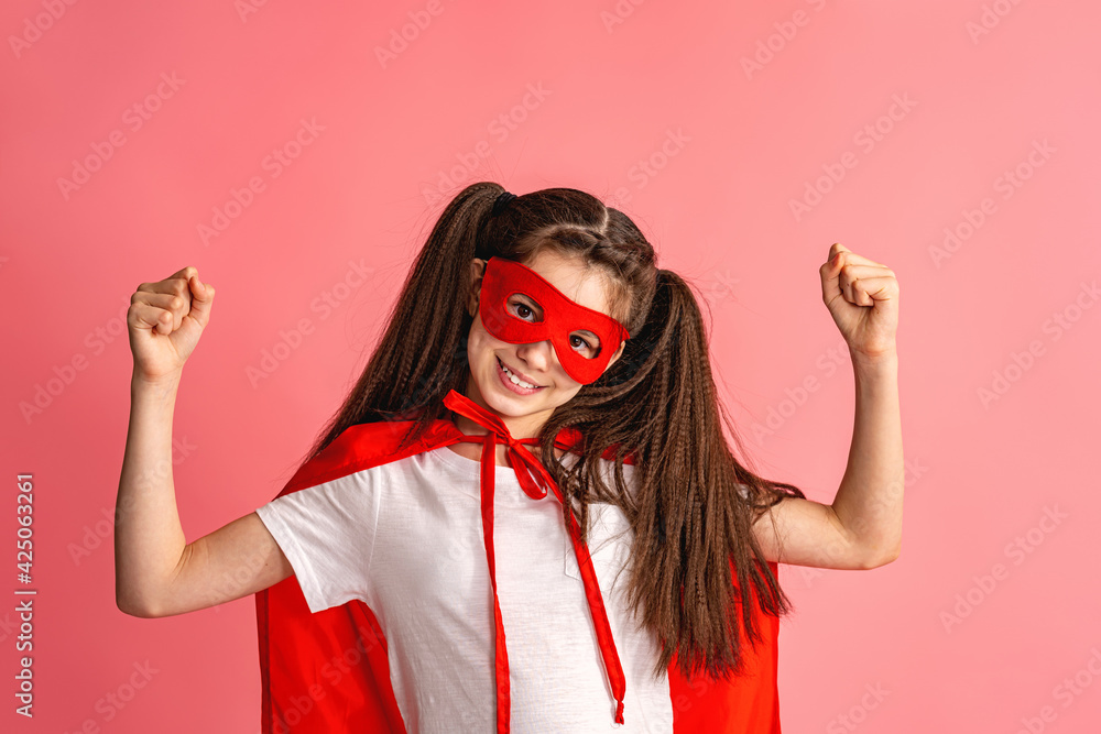 Fototapeta premium beautiful smiling girl in superhero costume, in red cape and mask shows how strong she is, isolated on pink background. Cute baby playing superhero game. concept of Girl Power. Copy the space.