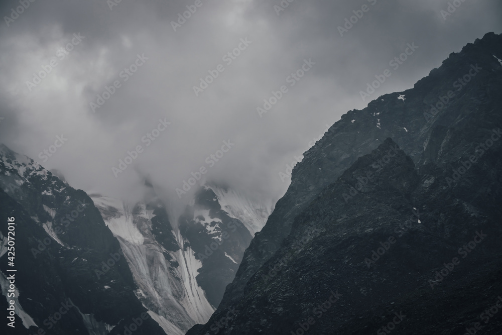 Dark atmospheric mountain landscape with glacier on black rocks in lead gray cloudy sky. Snowy mountains in low clouds in rainy weather. Gloomy landscape with black rocky mountains with snow in fog.