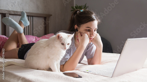 the girl is lying on the bed in front of an open laptop in her room and her white cat is sitting next to her