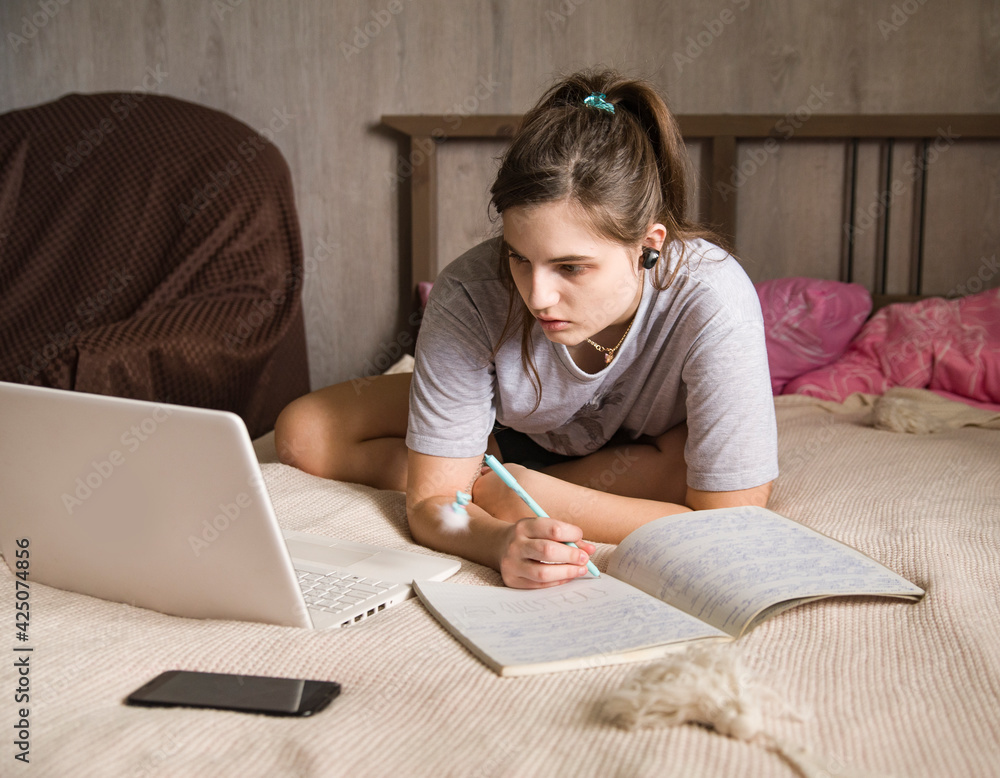 the girl sits on the bed in front of an open laptop in her room and writes something in a notebook the girl sits on the bed in front of an open laptop in her room and writes something in a notebook
