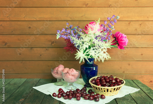 Beautiful bouquet in blue vase, glass bowls with cherry ice cream and basket of fresh cherry on rustic wooden table.