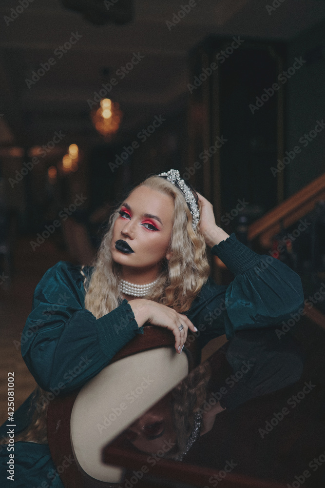 A blonde girl with a diadem in long hair and bright makeup posing on the stairs in the house.