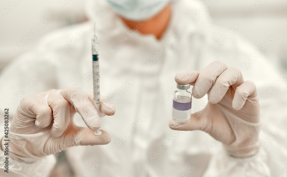 A doctor in a protective suit holds a syringe and an ampoule with the covid-19 coronavirus vaccine while standing in an intensive care unit in a modern hospital. A doctor in a protective suit holds a syringe and an ampoule with the covid-19 coronavirus vaccine while standing in an intensive care unit in a modern hospital.