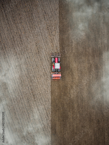 Red tractor plowing farm field aerial view looking down from above through clouds. Drone photo  cultivation machine equipment working ploughing countryside arable soil driven by farmer spring planting