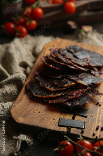 Delicious smoked sausage on a dark wooden board with tomatoes and spices. Delicious handmade snack made from natural ingredients. Pieces of dry pork, beef, chicken snacks. Meat products. Close-up.