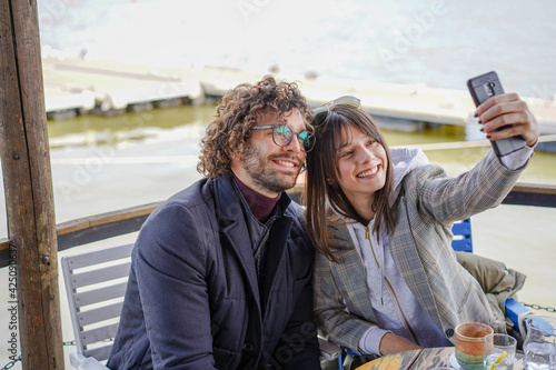 Beautiful young couple siting on river bank smiling and enjoying life. Young guy wearing glasses and have curly hair and beard. Girl is holding mobile phone and taking selfie. 