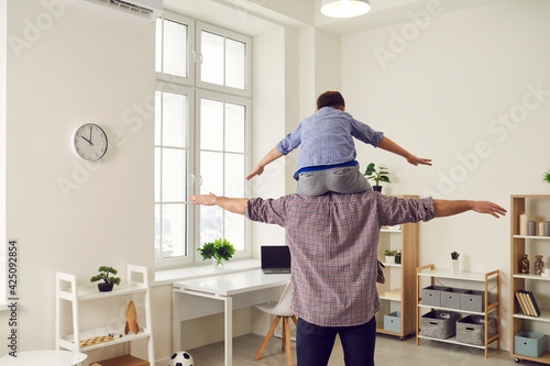 Fathers day. Little boy sits on his dad's shoulders and they have fun imitating a flight. Father at home plays with his son by placing him on his shoulder and standing with his back to the camera.