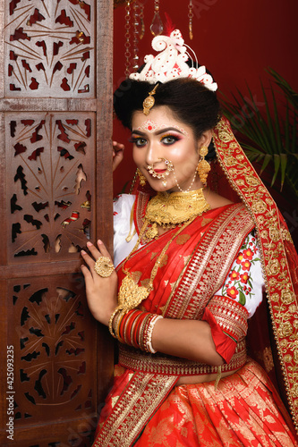 Stunning Indian bride dressed in traditional red Banarasi saree with heavy gold jewellery and veil smiles tenderly in studio lighting. Ethnic Bengali wedding.