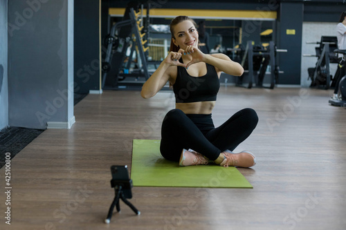 Healthy woman workout on mobile phone, Female sports blogger recording video on smartphone. Girl showing heart sign. Online exercise lesson at home.