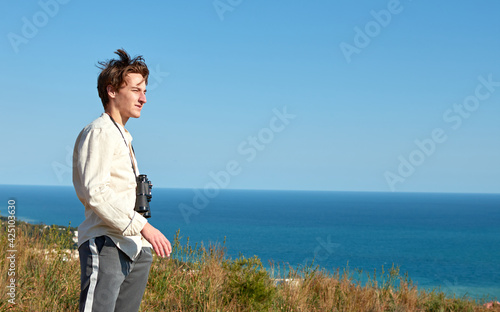 A portrait of a tired young man with binoculars hanging around his neck standing in front of lovely sea scenery looking afar
