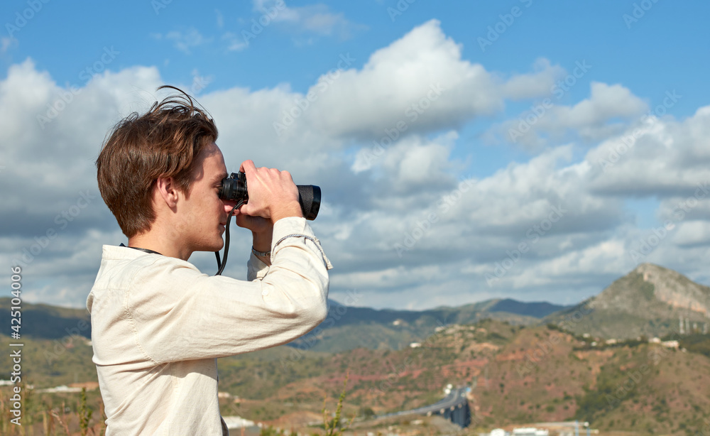 A side view of a young traveler from Spain in a white shirt looking over mountains through binoculars