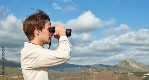 A side view of a young traveler from Spain in a white shirt looking over mountains through binoculars