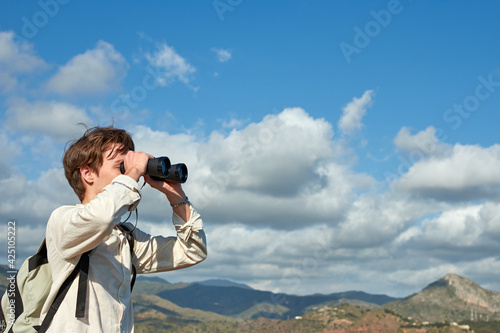 A shallow focus shot of a young traveler from Spain in a white shirt looking over mountains through binoculars
