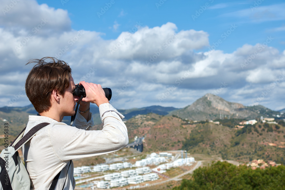 A Shallow focus shot of a young traveler from Spain in a white shirt looking over mountains through binoculars