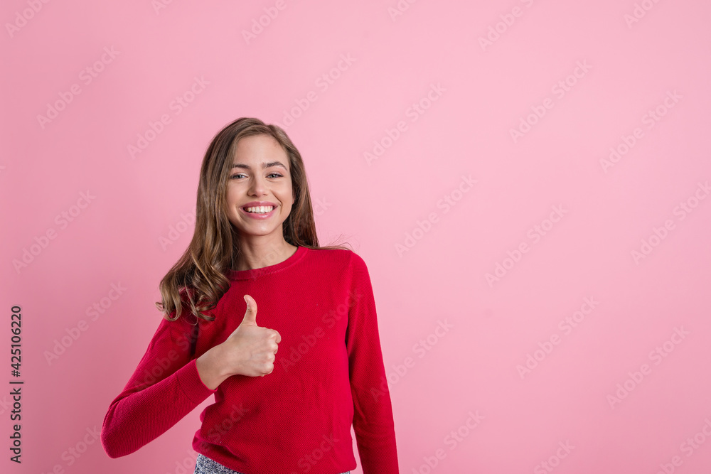 A beautiful young blonde girl in a red sweatshirt shows her thumbs up, a sign of approval on a pink background to copy the space. positive beautiful girl smiles and shows super A beautiful young blonde girl in a red sweatshirt shows her thumbs up, a sign of approval on a pink background to copy the space. positive beautiful girl smiles and shows super