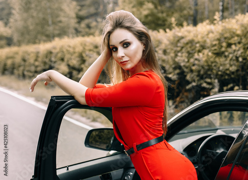 A beautiful young girl in a red overalls stands by a black car on an empty road in the forest © Daria Lukoiko